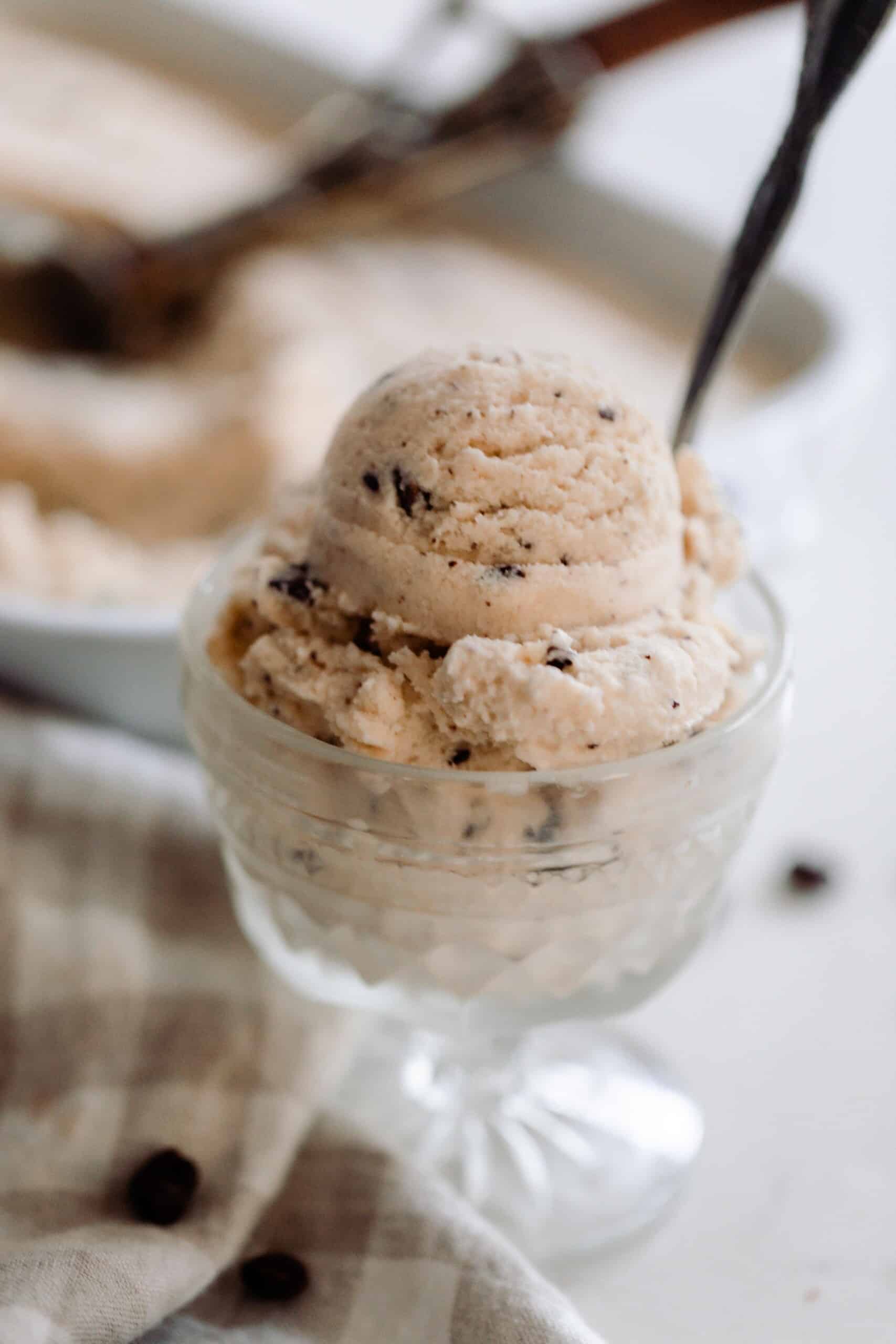 java chip ice cream scooped into a glass vintage ice cream dish with a spoon. The dish sits on a a white countertop op with a tan and white checked towel