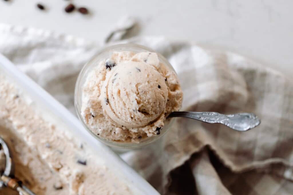 overhead photo of coffee chocolate chip ice cream in a ice cream dish on a tan and white checked towel with a baking dish of ice cream in front