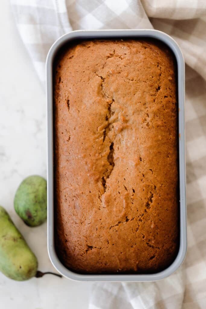 overhead photo of pawpaw bread in a loaf pan on a white and tan checked towel with two pawpaws to the left