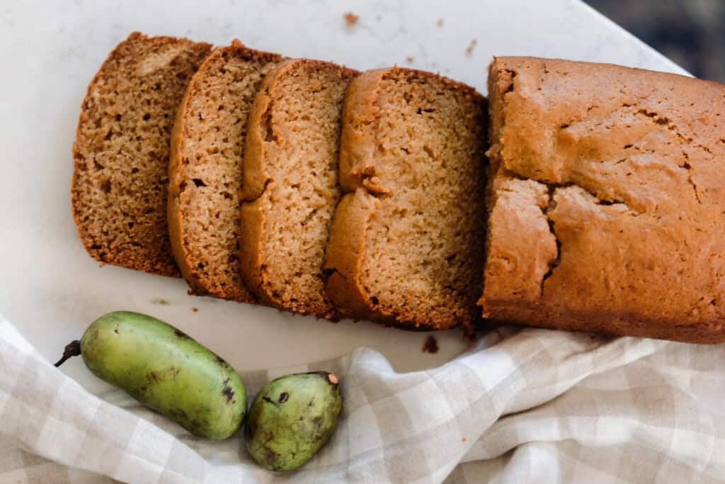 overhead photo of a loaf of pawpaw bread with four slices sliced onto a countertop. Two fresh pawpaws are on a white and tan checked towel