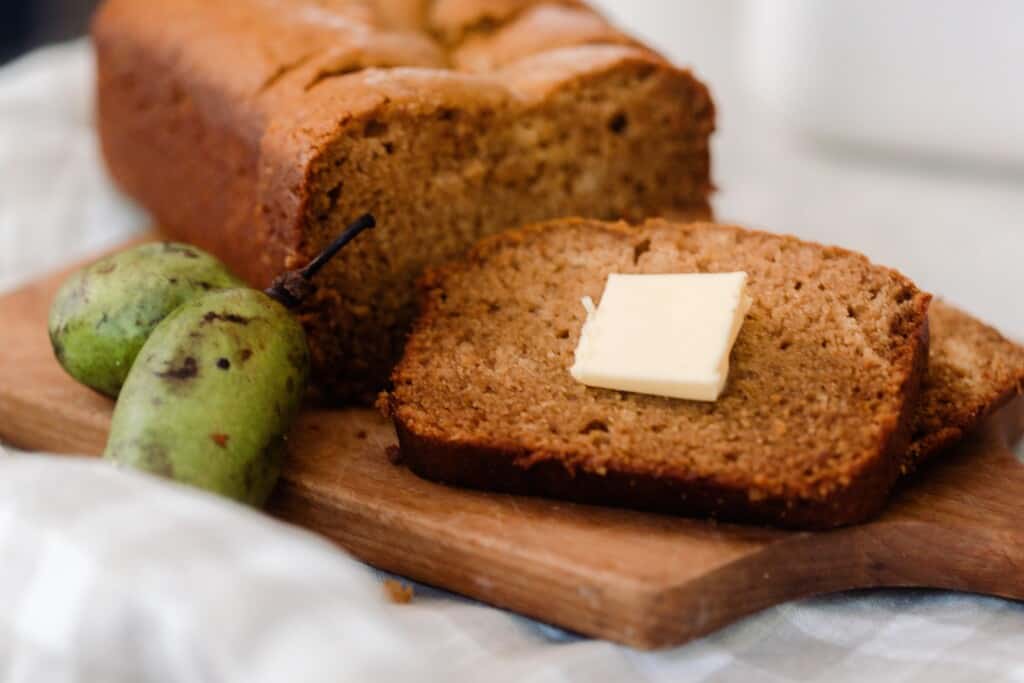 pawpaw bread on a cutting board with one slice off topped with butter. Two pawpaws sit on the cutting board