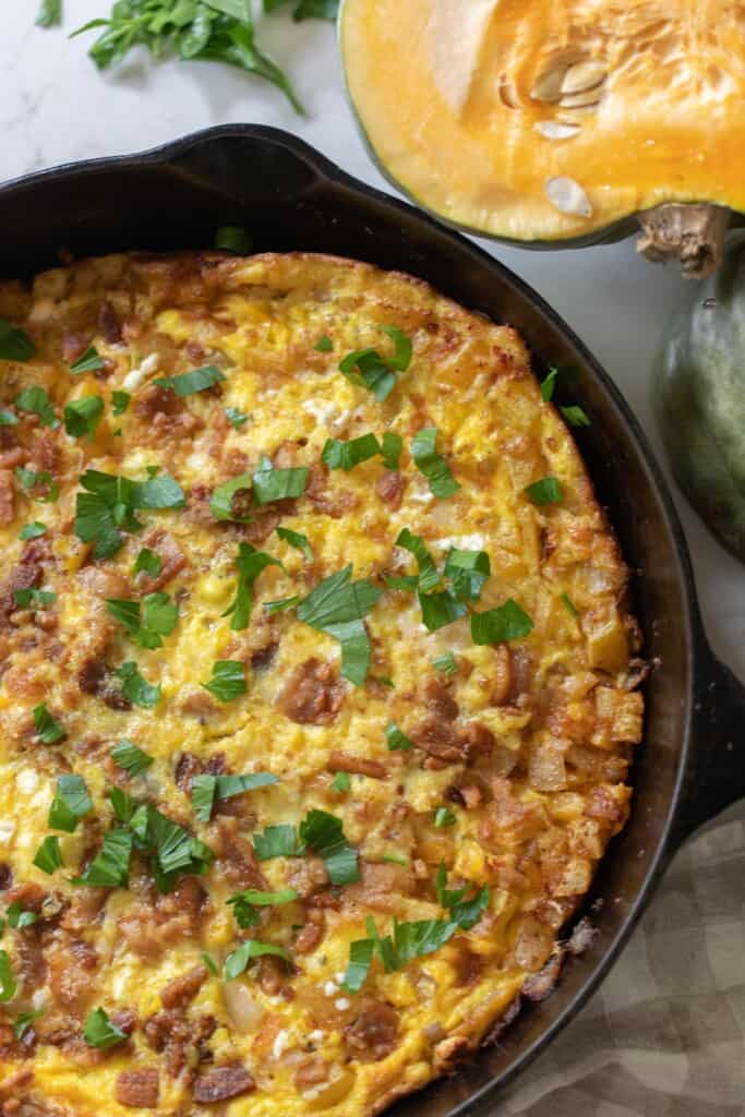 overhead photo of a pumpkin frittata topped with cheese and parsley in a cast iron skillet with a pumpkin sliced in half in the back right corner