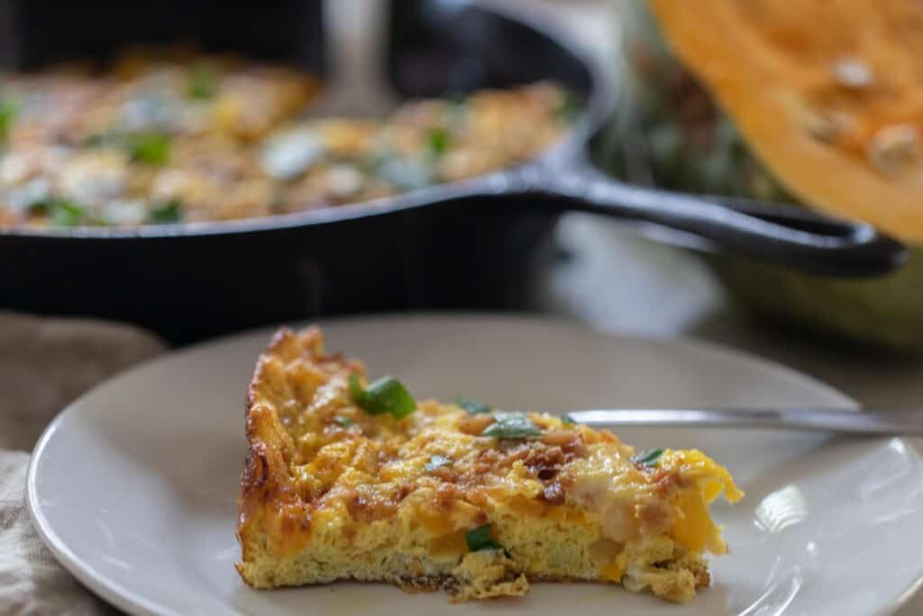 slice of pumpkin frittata on a white plate with a cast iron skillet of the remaining egg dish and a half of pumpkin in the background