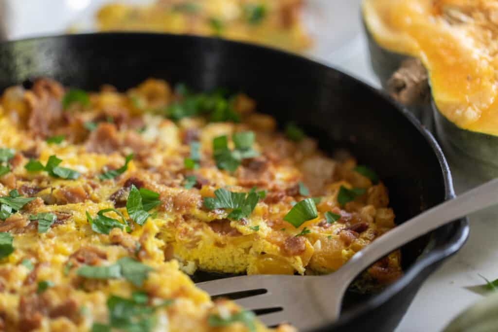 side view of a pumpkin frittata topped with feta, cheddar cheese and fresh herbs in a cast iron skillet with a halved pumpkin in the background