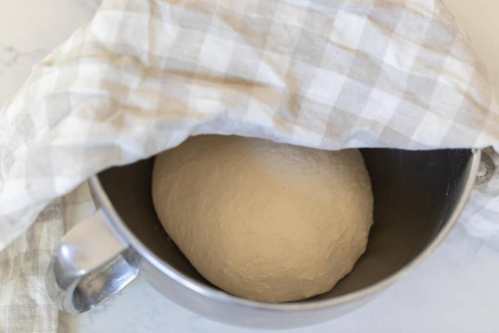dough in a stainless bowl with a tan and white checked towel covering half of the bowl