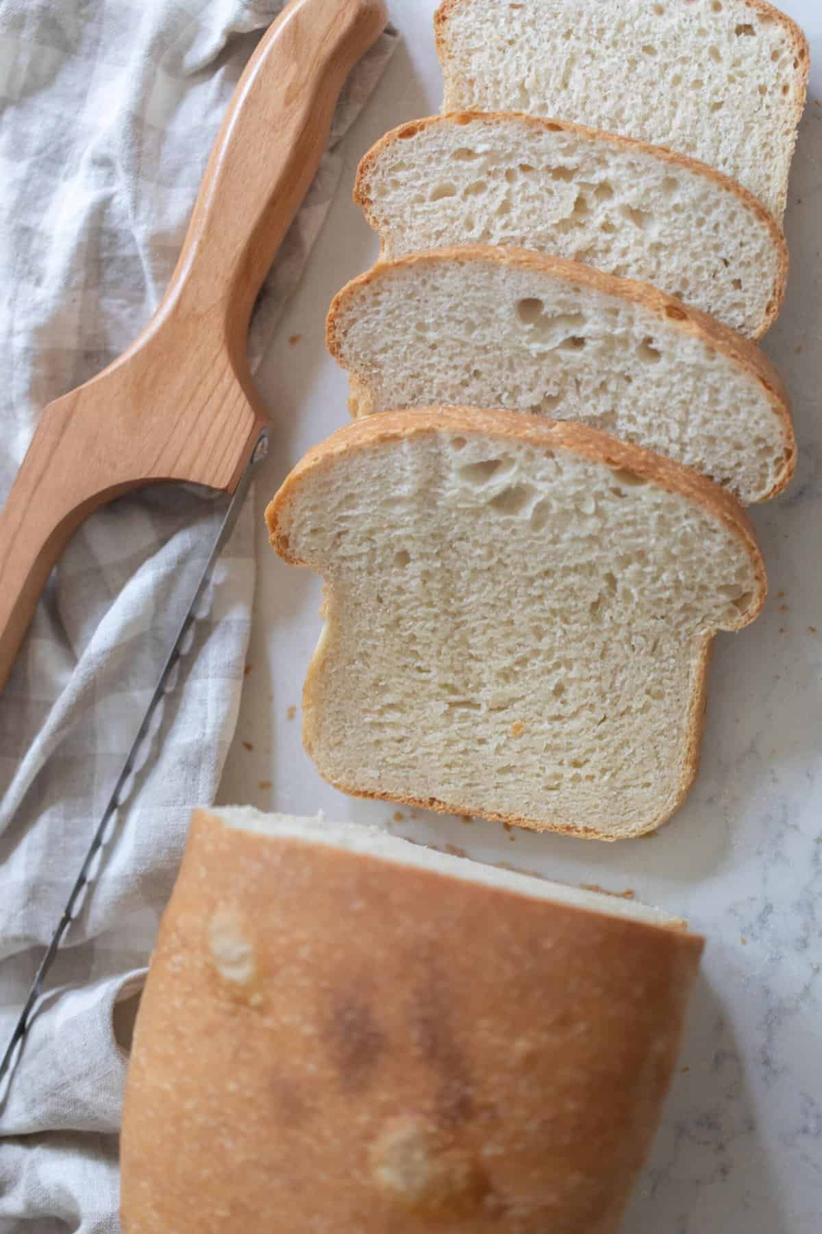 overhead photo of a loaf of sourdough sandwich bread with four slices off on white countertop with a towel and bread knife to the left