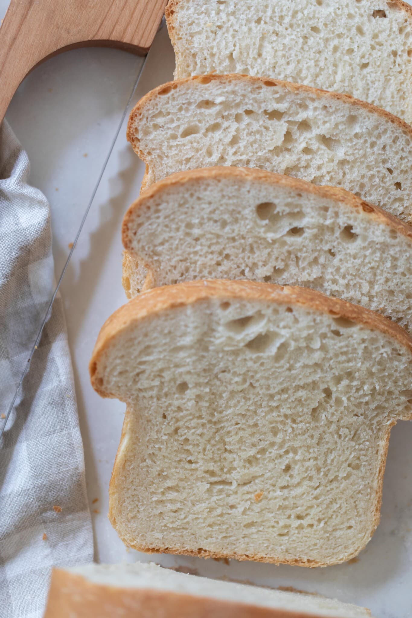 overhead photo of four slices of soft sourdough discard sandwich bread on a white countertop with a towel and wood knife to the left