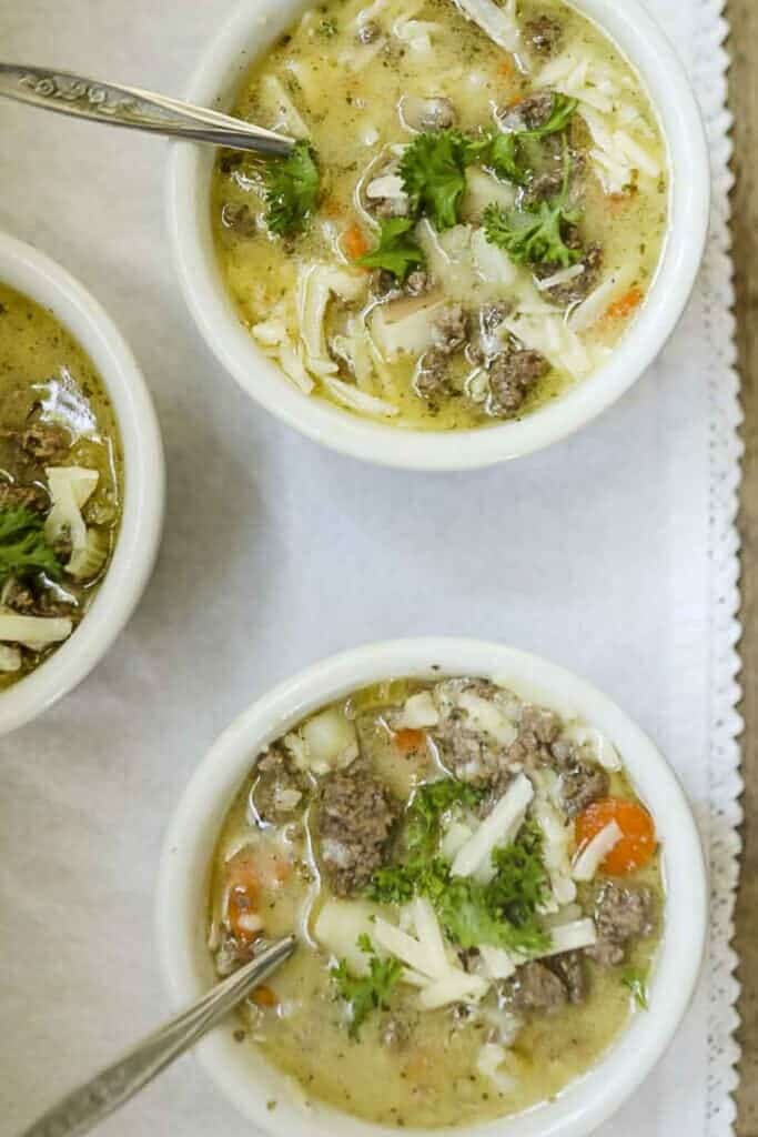 overhead photo of three bowls of cheeseburger soup topped with cheese and fresh herbs on a white table runner