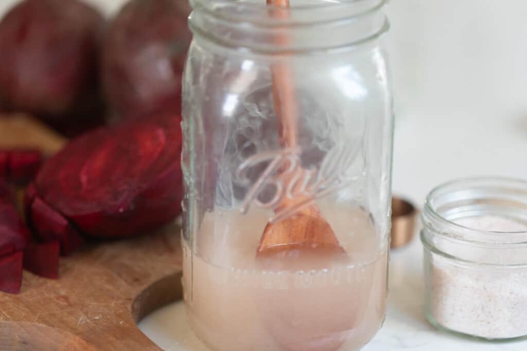 salt being dissolving in water in a jar with a wooden spoon on a white countertop with a cutting board of beets to the left