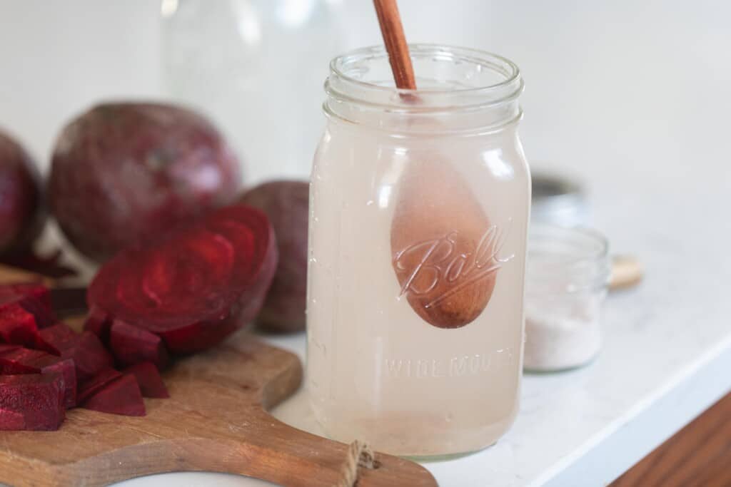 quart size mason jar filled with salt water with a wooden spoon on a white countertop. Beets are on a cutting board to the left