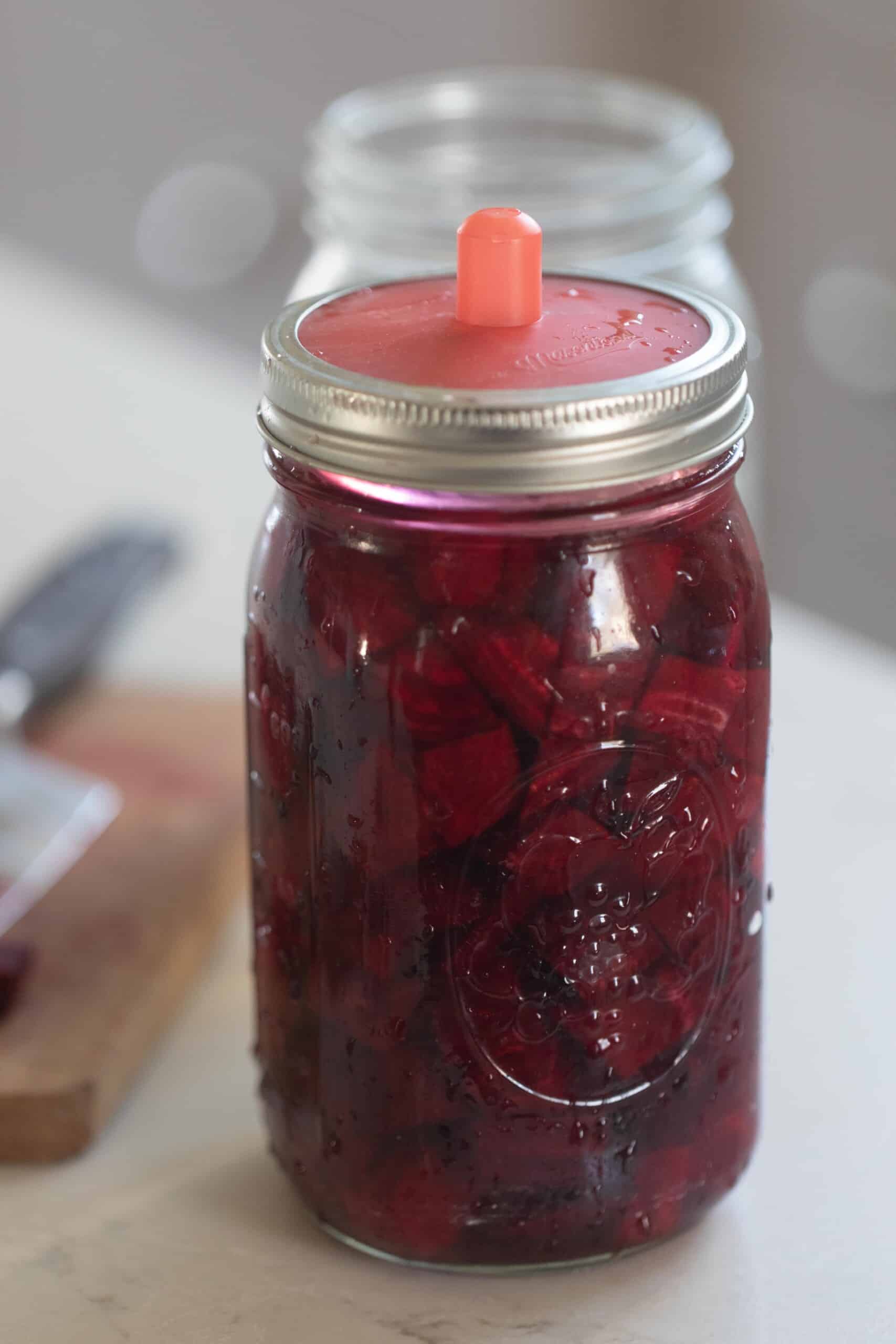 fermented beets in a glass mason jars with a silicon fermenting lid on a white countertop with a cutting board to the left