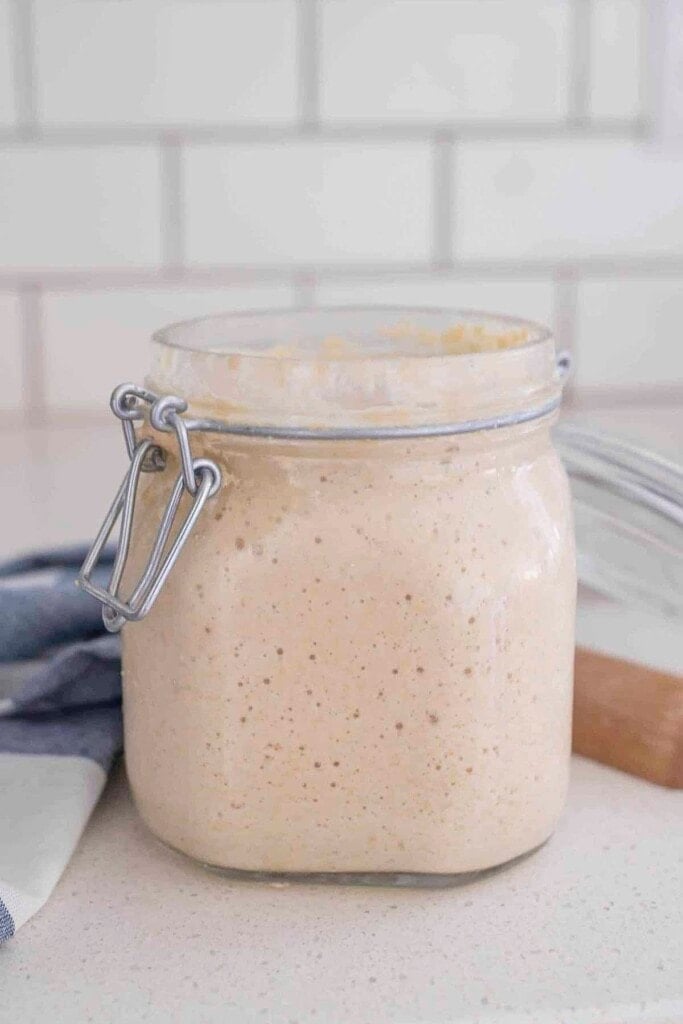 sourdough starter in a glass jar with a swing top lid on a white countertop with a wooden spoon to the right 
