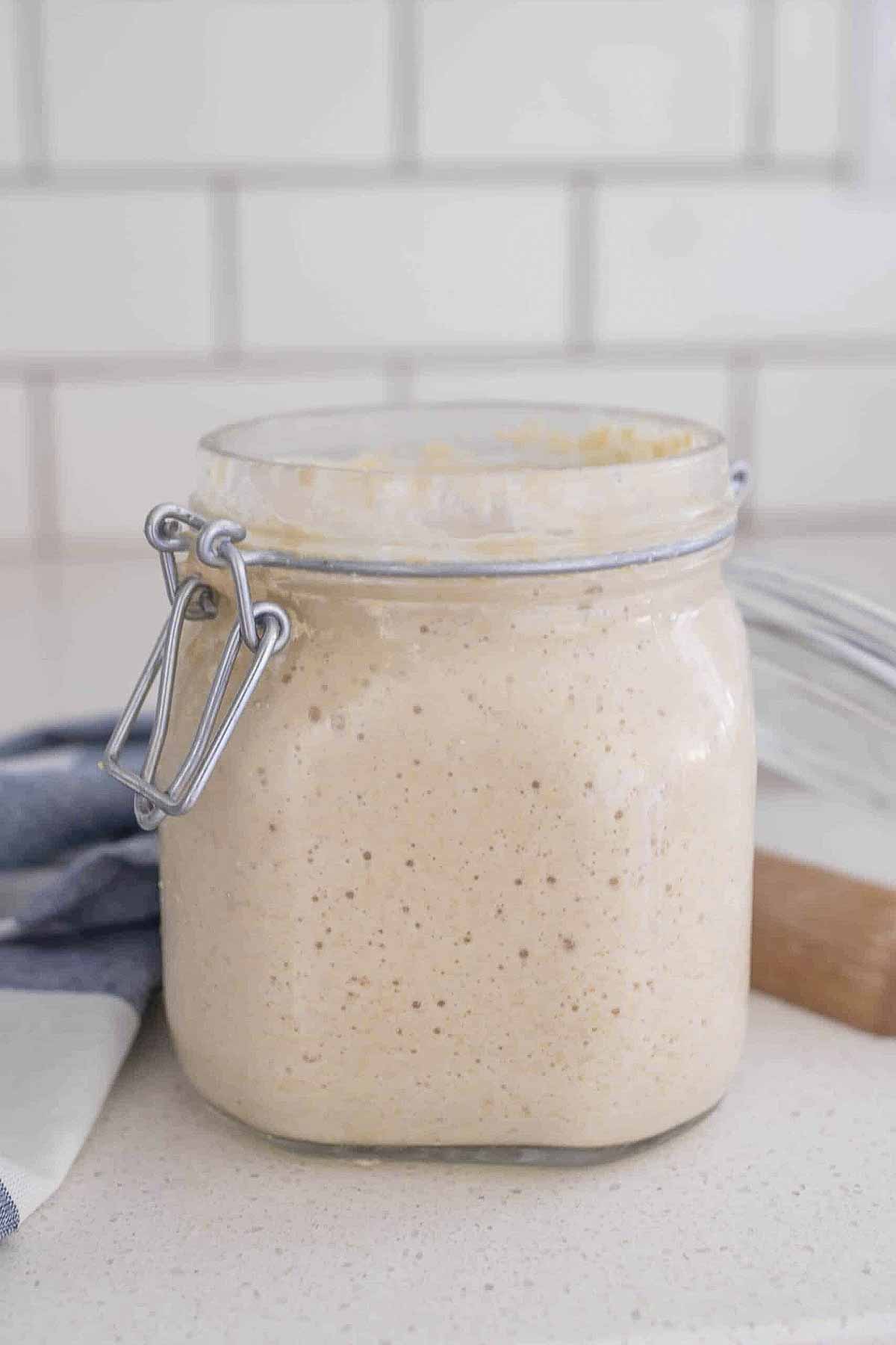 sourdough starter in a glass jar with a swing top lid on a white countertop with a wooden spoon to the right.