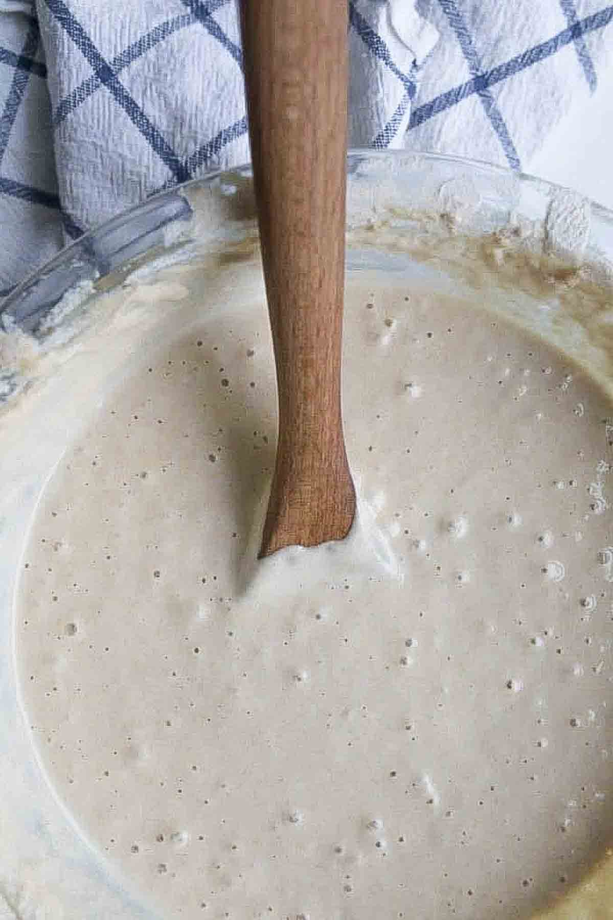 overhead photo of a sourdough starter in a glass bowl with a wooden spoon in the starter. The bowl is next to a white and blue checked towel