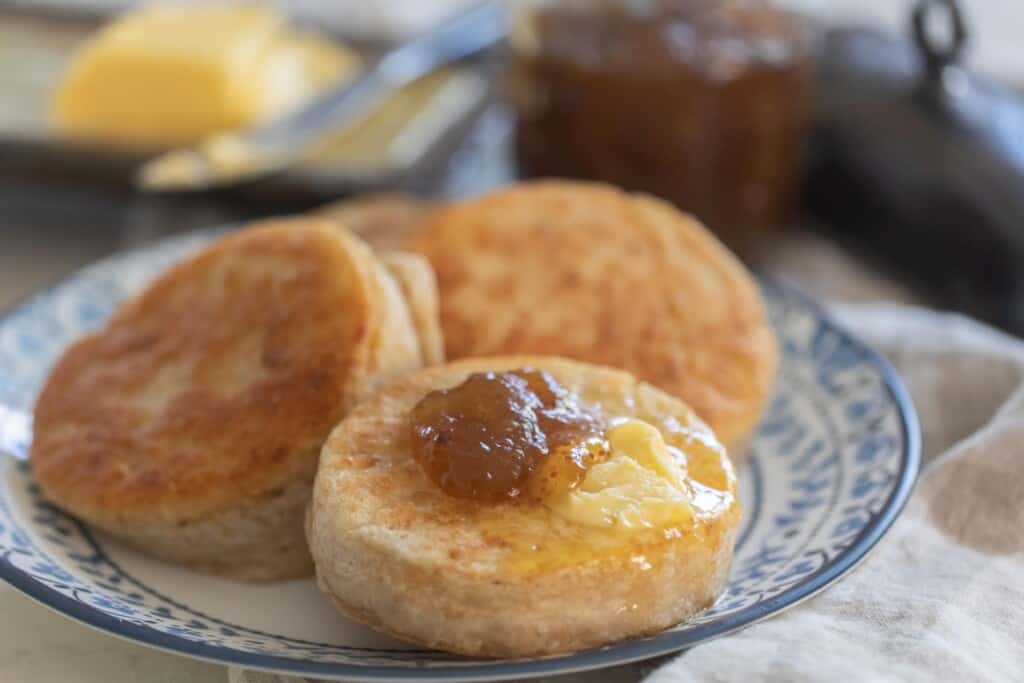 three sourdough crumpets on a blue and white plate topped with butter and jam with more butter and jam in the background