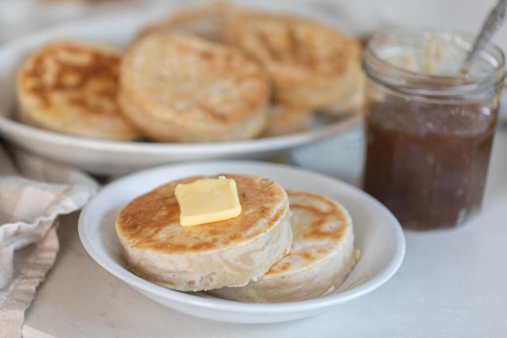 two sourdough crumpets topped with butter on a white plate with a jar of jam and a plate of more crumpets in the background