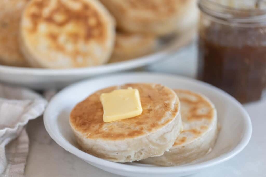 two sourdough crumpets topped with butter on a white plate with a platter of more crumpets and a jar of jam in the background