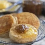 three sourdough crumpets on a blue and white plate topped with butter and jam with more butter and jam in the background