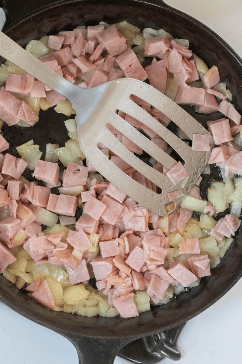 Cubes of ham added to the caramelized onions in a skillet with a spatula.