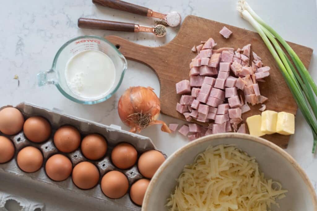 Ingredients for ham and cheese frittata laid out with a cutting board on a white countertop