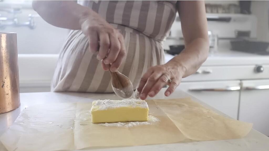 two stick of butter on parchment paper. A woman adding flour to the top of the butter