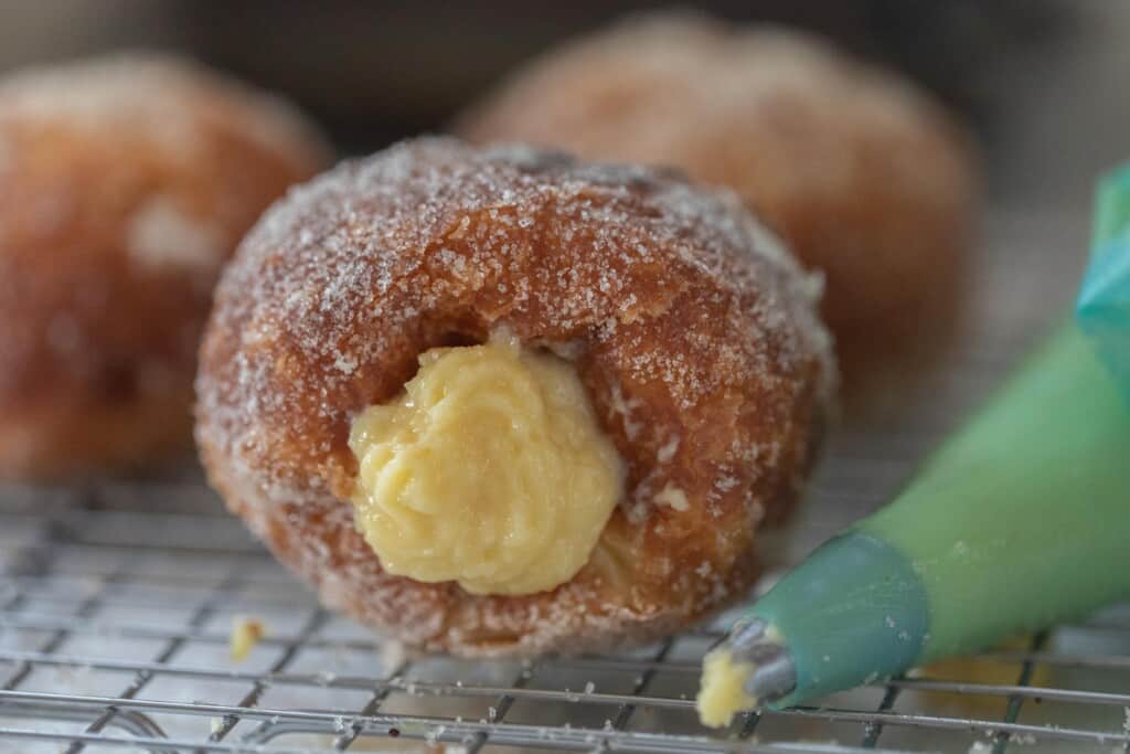 Brioche donut with custard filling with a green piping bag next to it on a wire rack