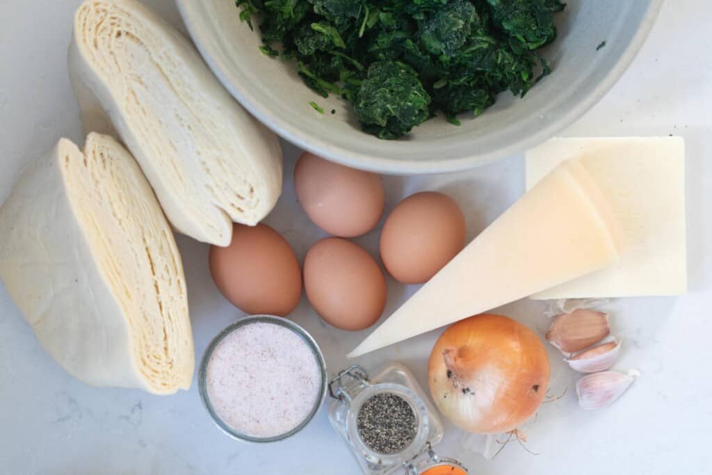 overhead photo of eggs, puff pastry, onion, salt, spinach and salt on a white countertop