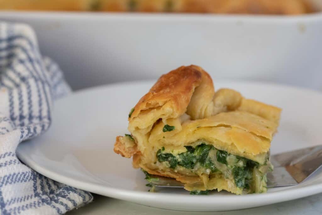 a slice of spinach pie made with puff pastry on a white plate with a blue and white stripped towel to the left