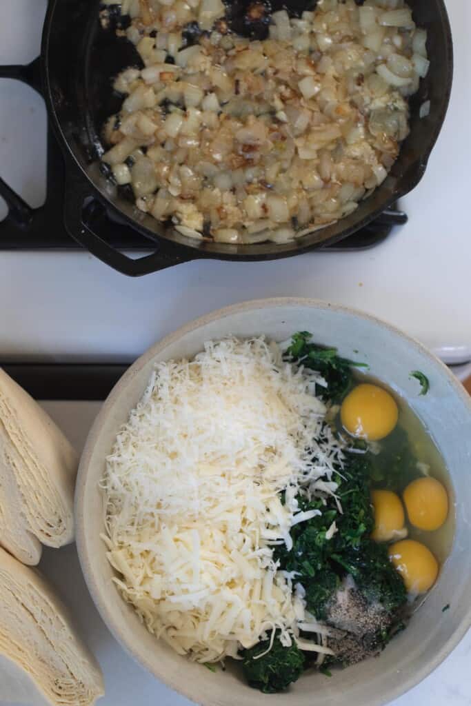 spinach, cheese, and eggs in a bowl with a cast iron skillet of onions behind the bowl