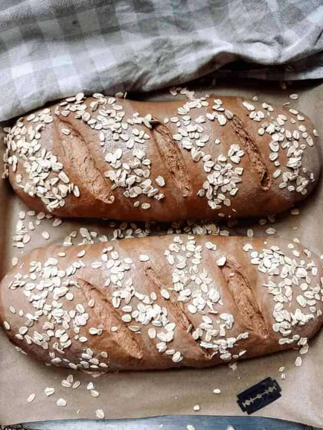 Two loaves of sourdough brown bread dough on parchment paper scored and sprinkled with rolled oats next to a checkered tea towel