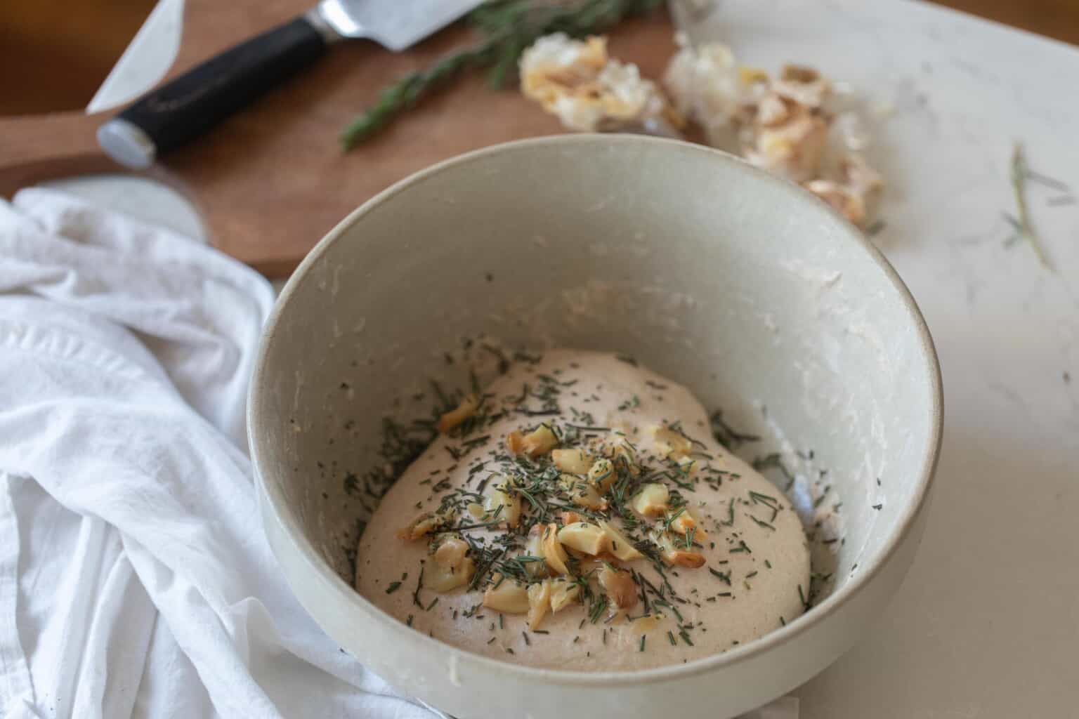 roasted garlic and rosemary added to the top of bread dough in a cream colored dish on a white countertop with white towel to the left and garlic in the background
