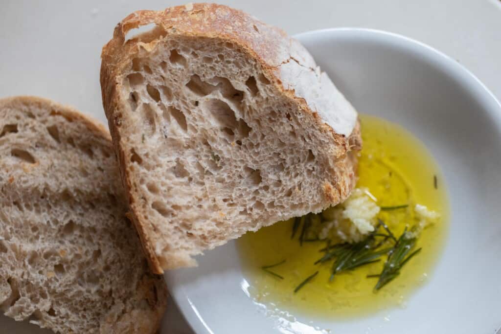 slice of sourdough garlic bread resting on a white bowl full of garlic, oil, and rosemary