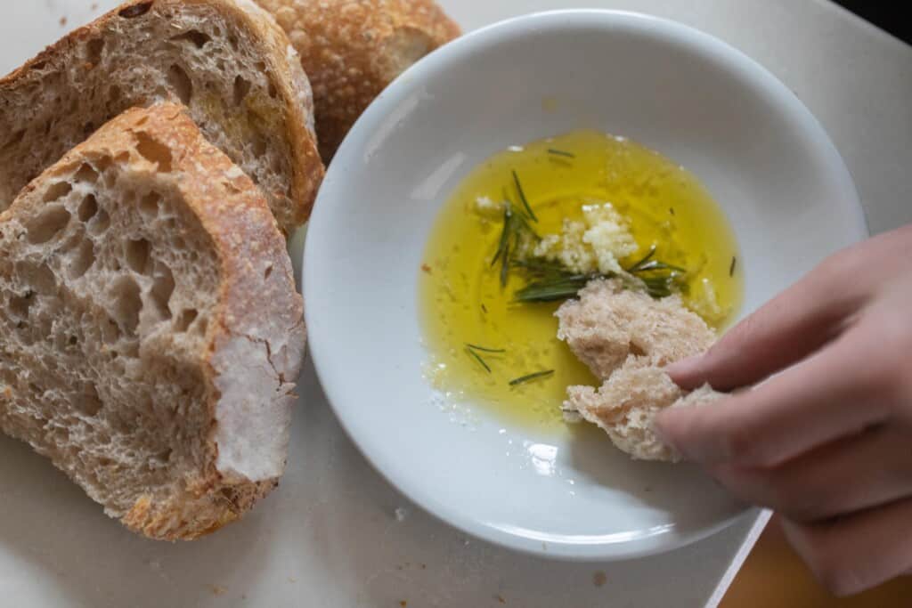 hand dipping sourdough bread into a bowl of oil and herbs with more bread surrounding the herbs