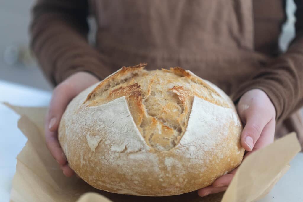 Woman holding a boule of high hydration sourdough with parchment paper underneath it. 