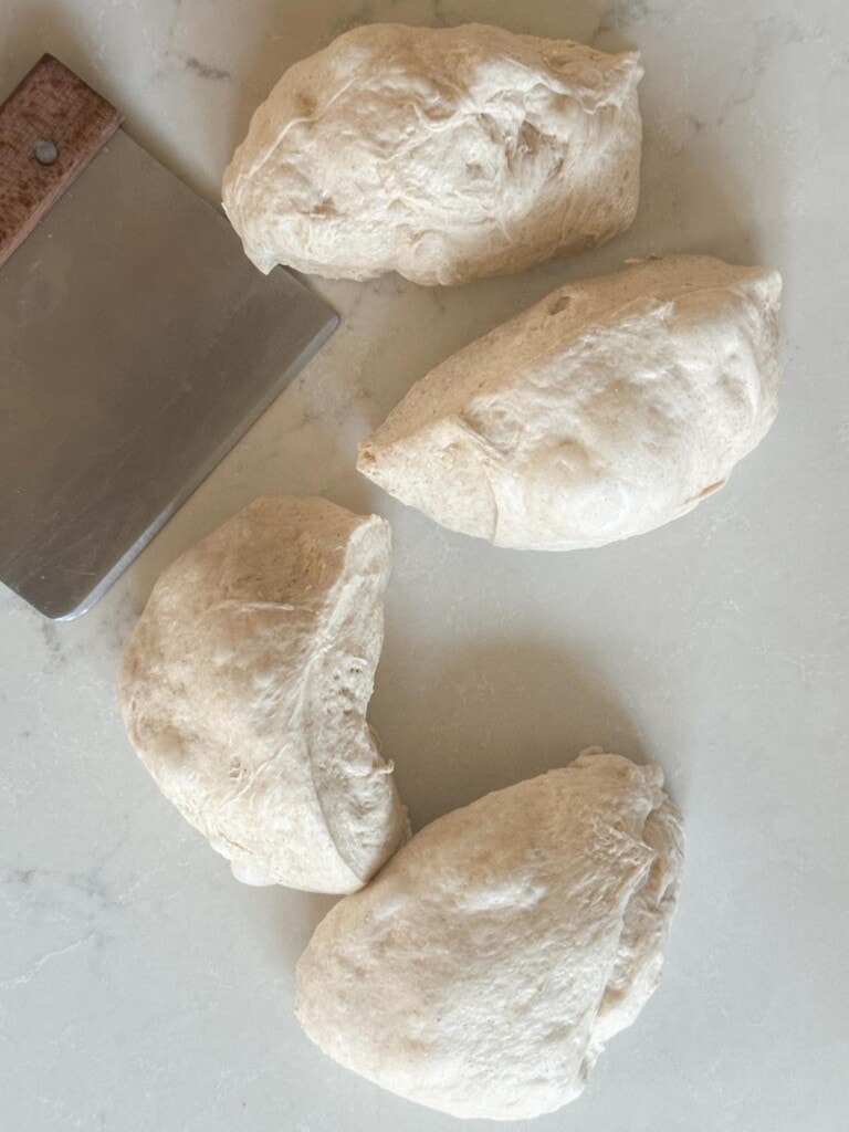 Mini sourdough loaf dough cut into 4 equal pieces with a bench scraper on the side on a white countertop