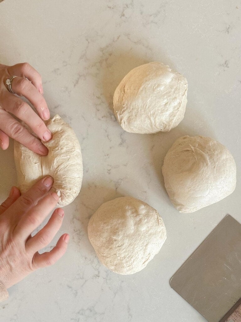 two hands shaping a dough ball with 3 other dough balls and a bench scraper on the side