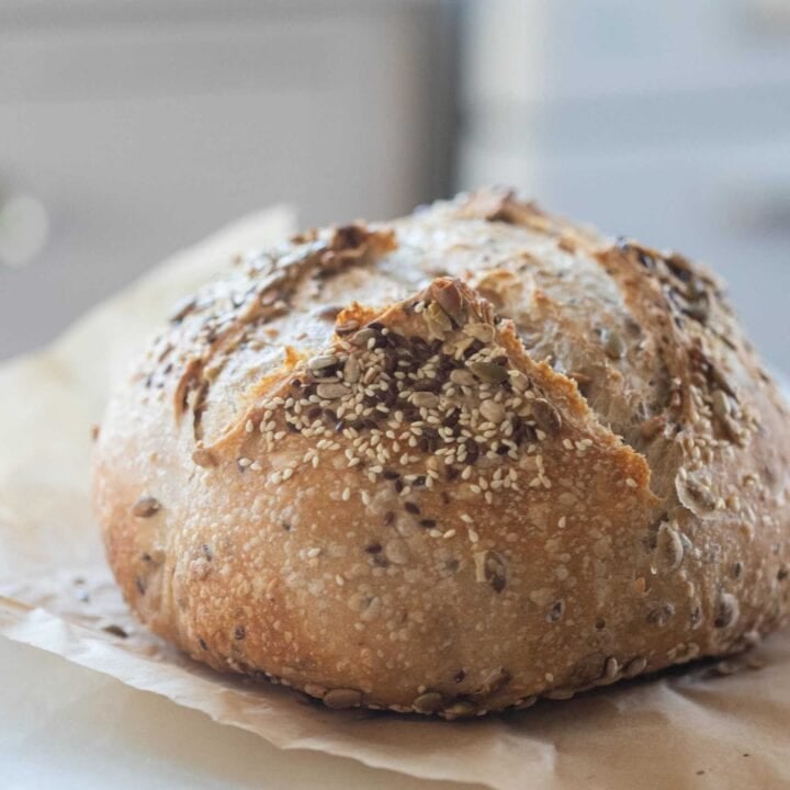 loaf of seeded sroudough bread on parchment paper with a white oven and cabinet int he background
