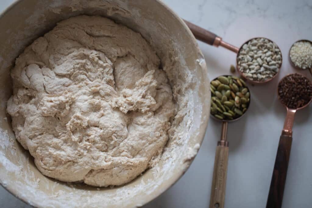 Seeded sourdough bread dough in a bowl with seeds in measuring cups on the side on a white countertop