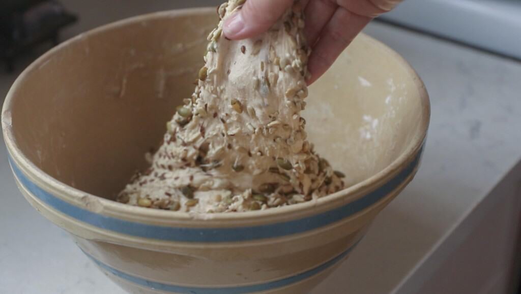 Hand stretching dough for seeded sourdough bread in a bowl on a white countertop