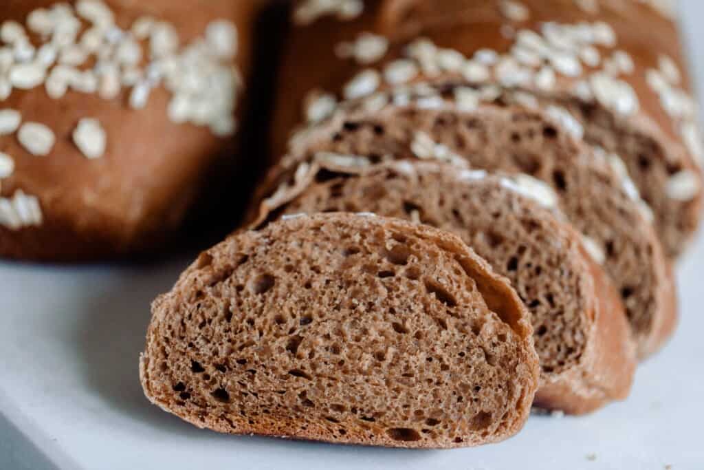 A loaf of sourdough brown bread sliced into with a slice facing the camera on a white countertop