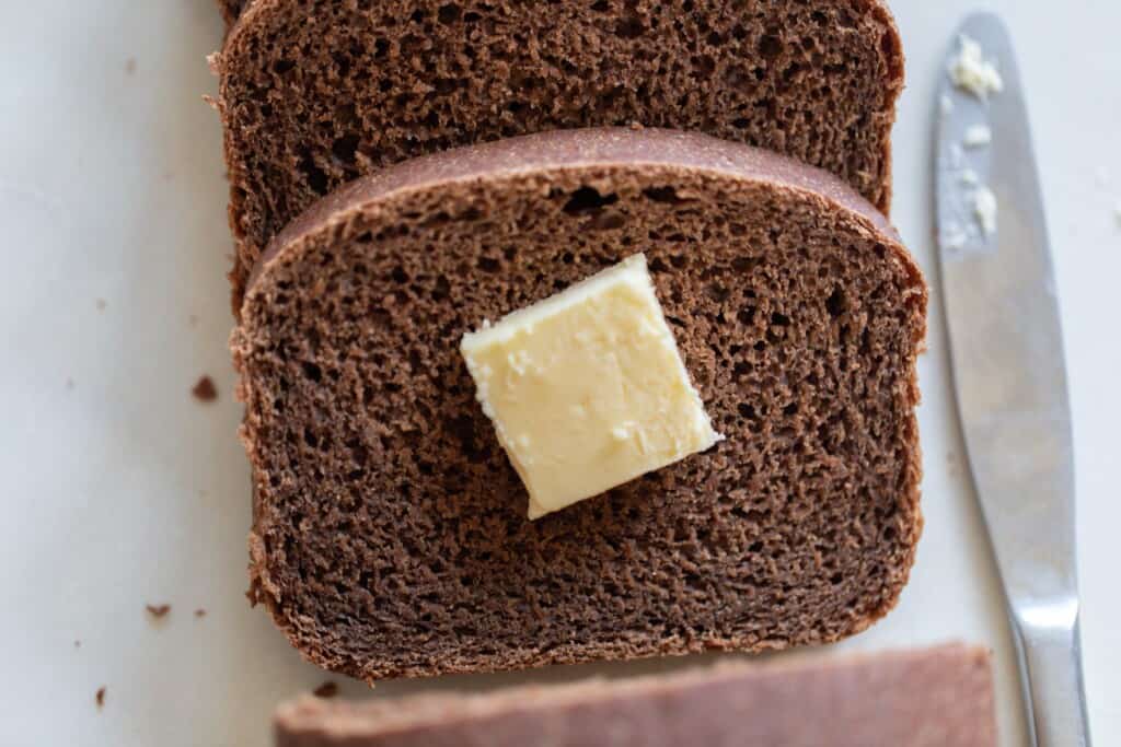 two slices on pumpernickel bread on a white countertop with the top slice of bread topped with a pat of butter on the top slice