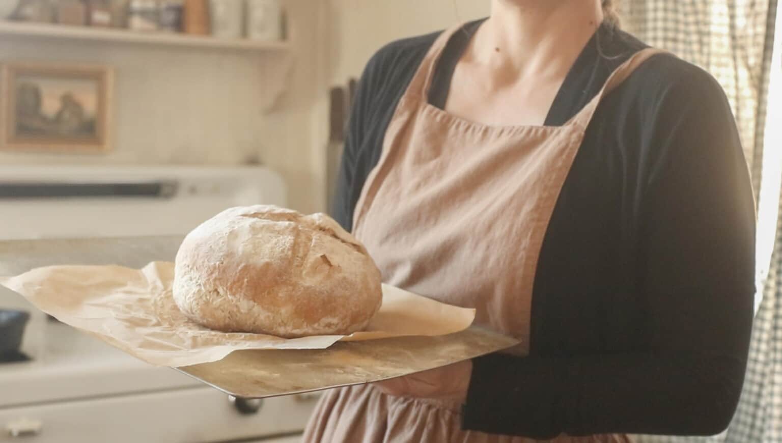 Woman in a black shirt and peach colored apron holding a cutting board with a piece of parchment paper under sourdough bread baked without a Dutch oven