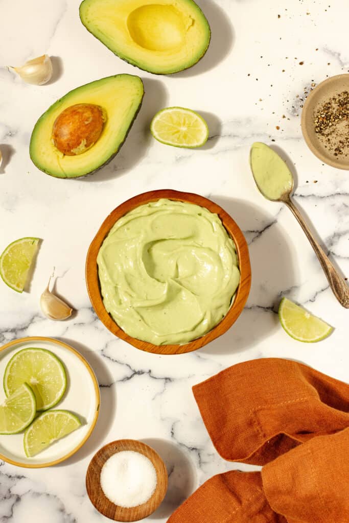 Overhead picture of avocado aioli in a terracotta bowl surrounding by the ingredients, including an avocado, lime, and salt