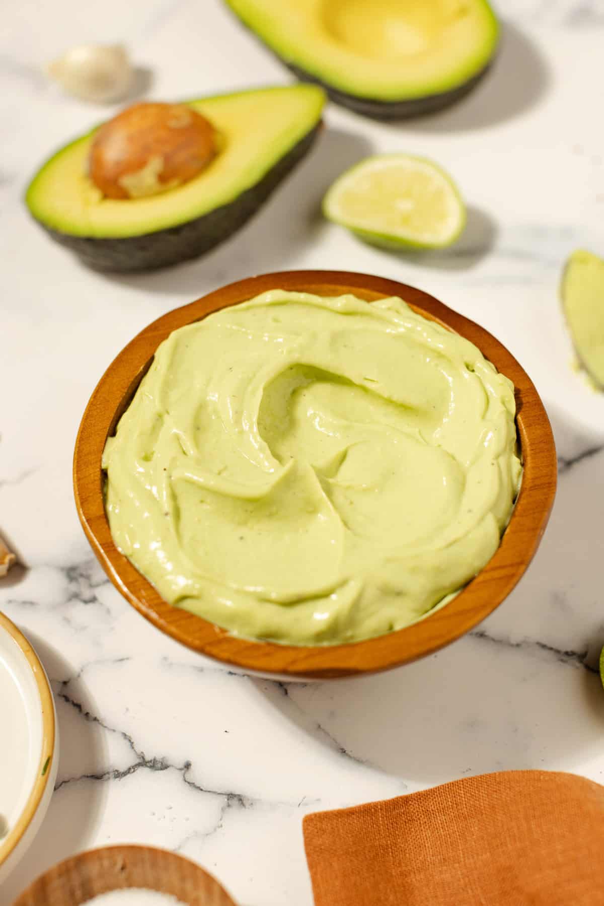 Avocado aioli in a terracotta bowl on a white countertop surrounded by ingredients, such as an open avocado and garlic