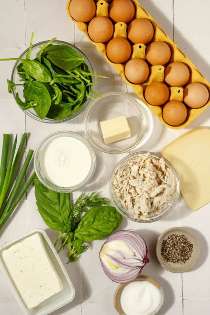 overhead view of chicken frittata ingredients including a dozen eggs, spinach, shredded chicken, milk, herbs, onion, salt, black pepper butter, and green onions