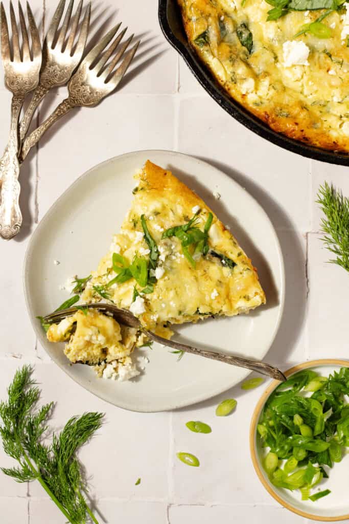 a slice of chicken frittata on a white plate with herbs and forks and a cast iron in the background