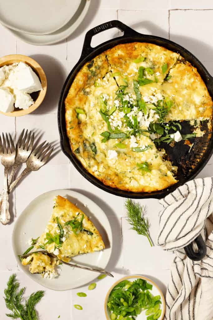 An overhead shot of a lice of frittata on a white plate next to a cast iron skillet full of the frittata on a white countertop