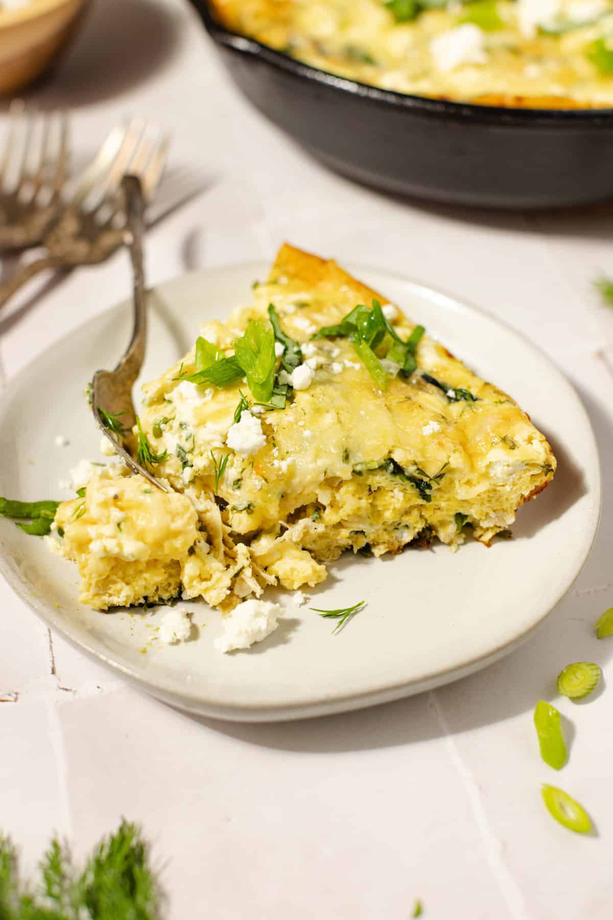 A piece of chicken frittata on a white plate with a fork and a cast iron skillet in the background