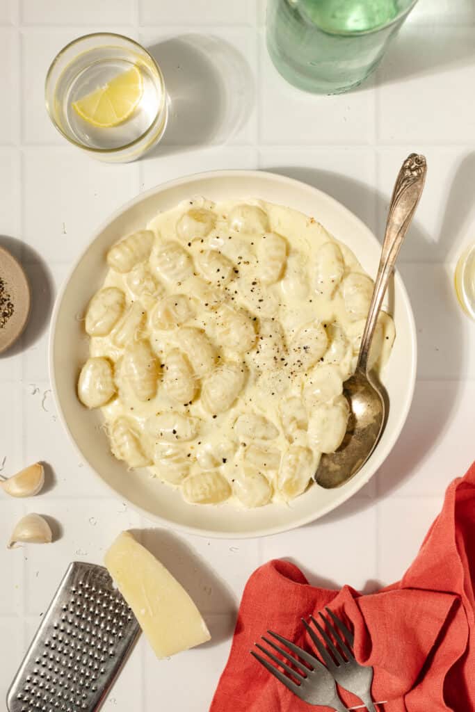 a cream colored plate with creamy sauce with gnocchi and a spoon. The bowl is surrounded by garlic clove, parmesan cheese, a microplane, red napkin, and glasses