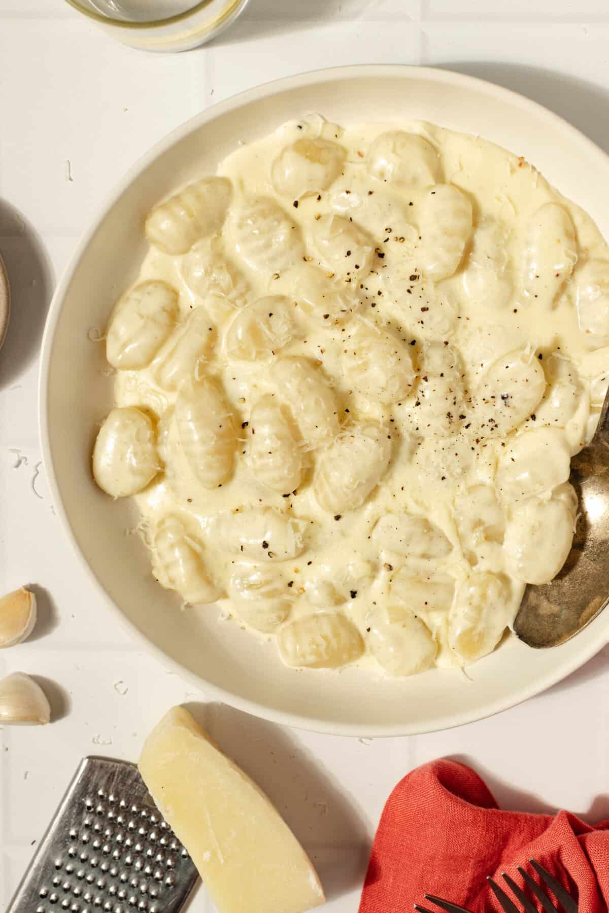 overhead photo of a bowl of gnocchi in a creamy sauce with a spoon. a red towel and ingredients surround the bowl